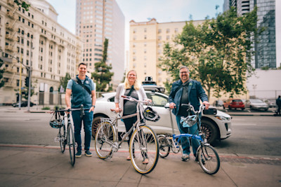 Ken McLeod, Caron Whitaker, and Bill Nesper standing on the sidewalk with bicycles in front of a parked Waymo vehicle